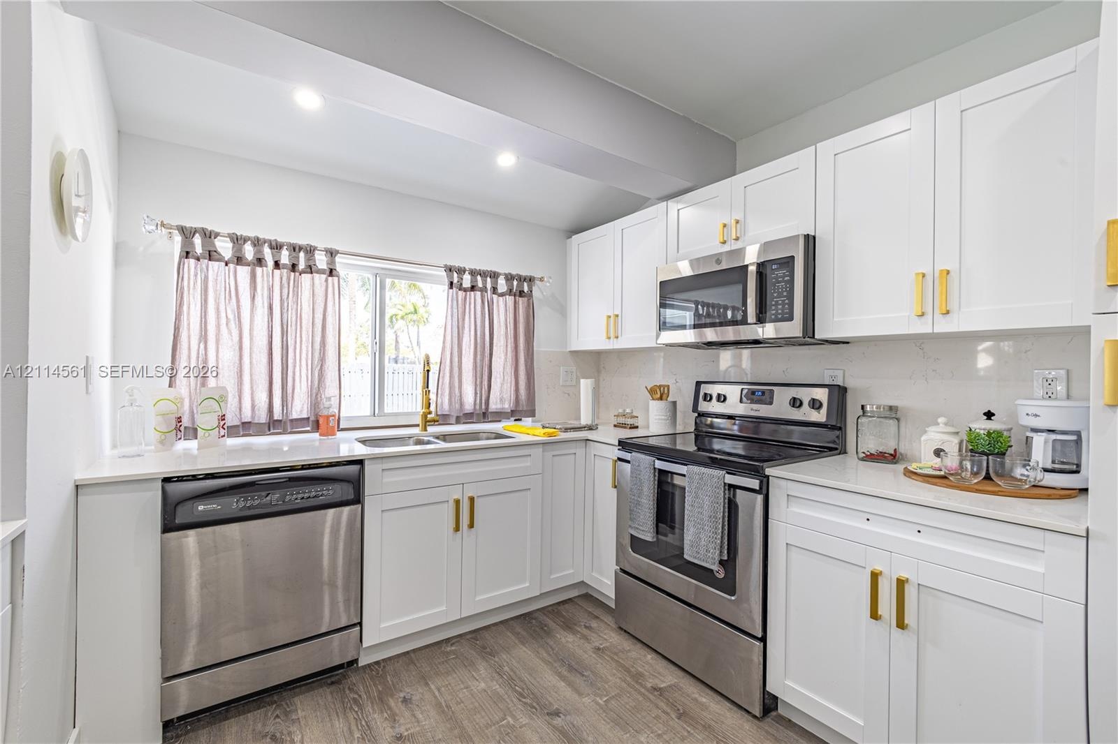Kitchen featuring white shaker-style cabinetry, metallic gold hardware, stainless steel appliances, light-toned stone-finish countertops, and wood-fin