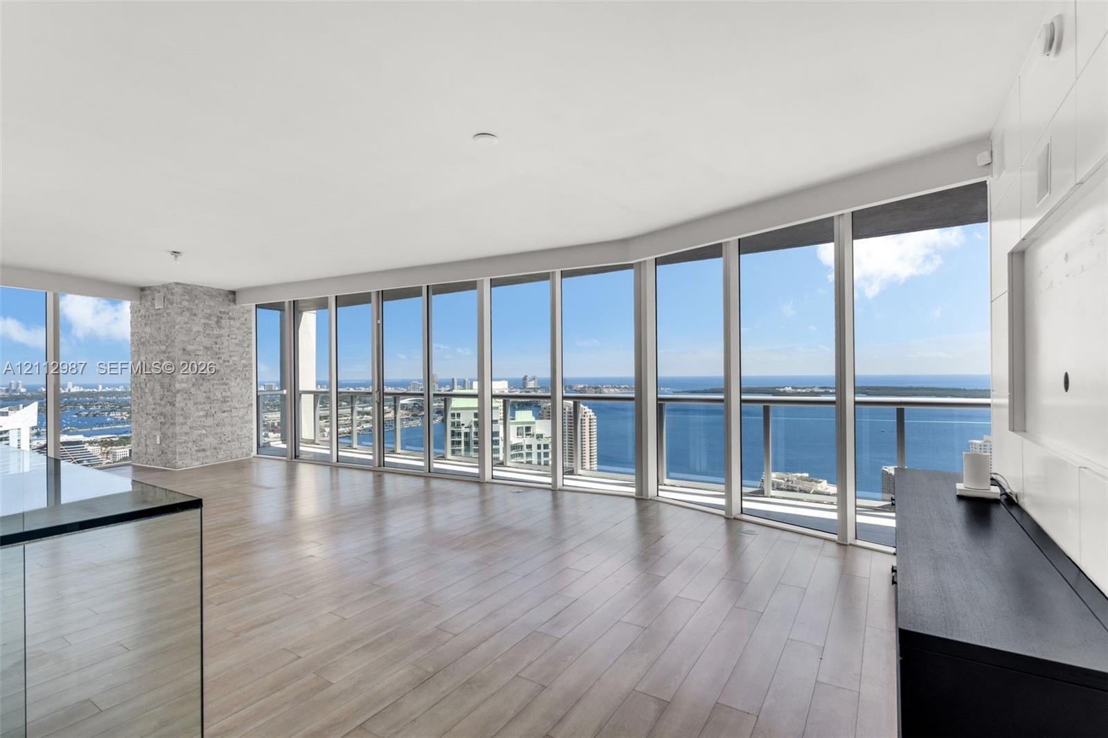 Floor-to-ceiling windows, wood-finish flooring, stone-veneer accent pillar, glass-panel balcony railing, and expansive water backdrop