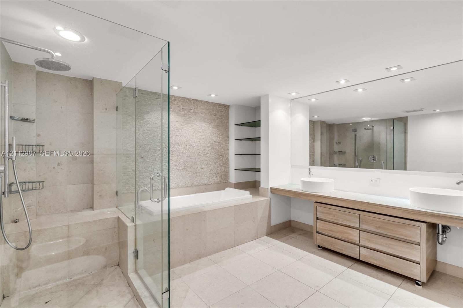 Floating wood-finish vanity with dual vessel sinks, glass-enclosed shower, built-in soaking tub, textured stone accent wall, and expansive frameless v
