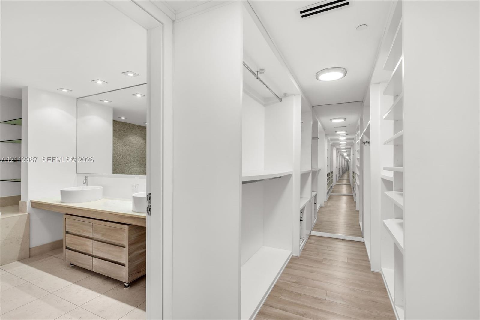 Walk-in closet featuring white built-in shelving, full-length mirror, and wood-finish flooring, accompanied by a bathroom with a dual-vessel sink vani