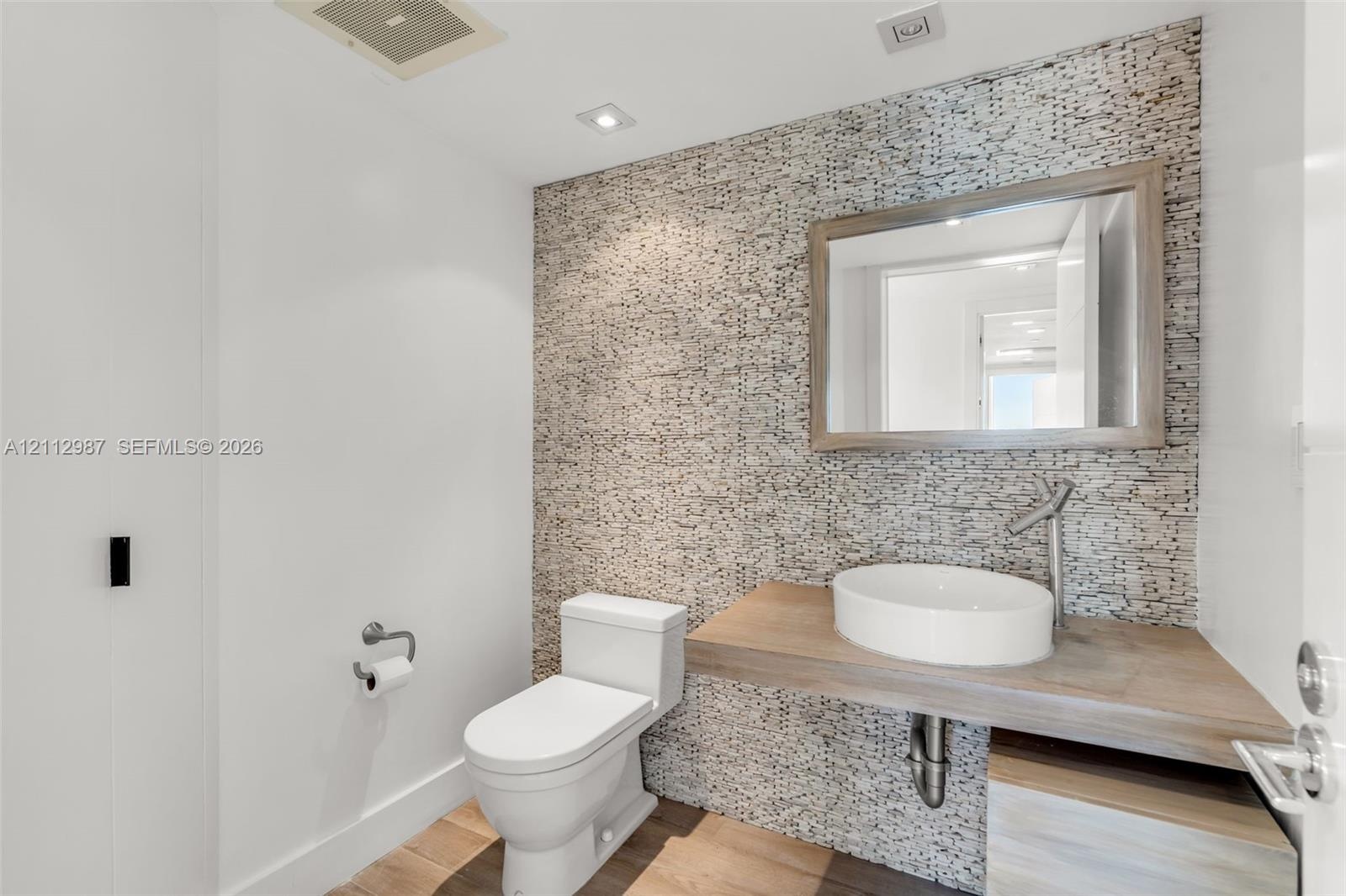 Textured stone accent wall featuring a wall-mounted wood-finish vanity, circular vessel sink, single-lever faucet, and wood-finish flooring