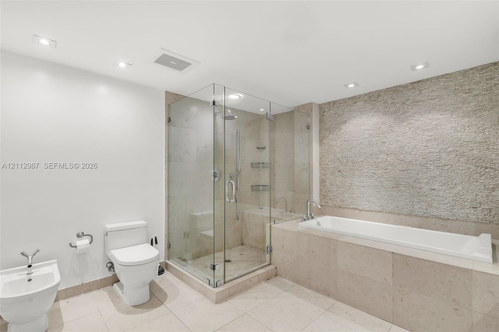 Bathroom featuring a frameless glass shower enclosure, a soaking tub with stone-tiled surround, a textured stone accent wall, recessed lighting, and a