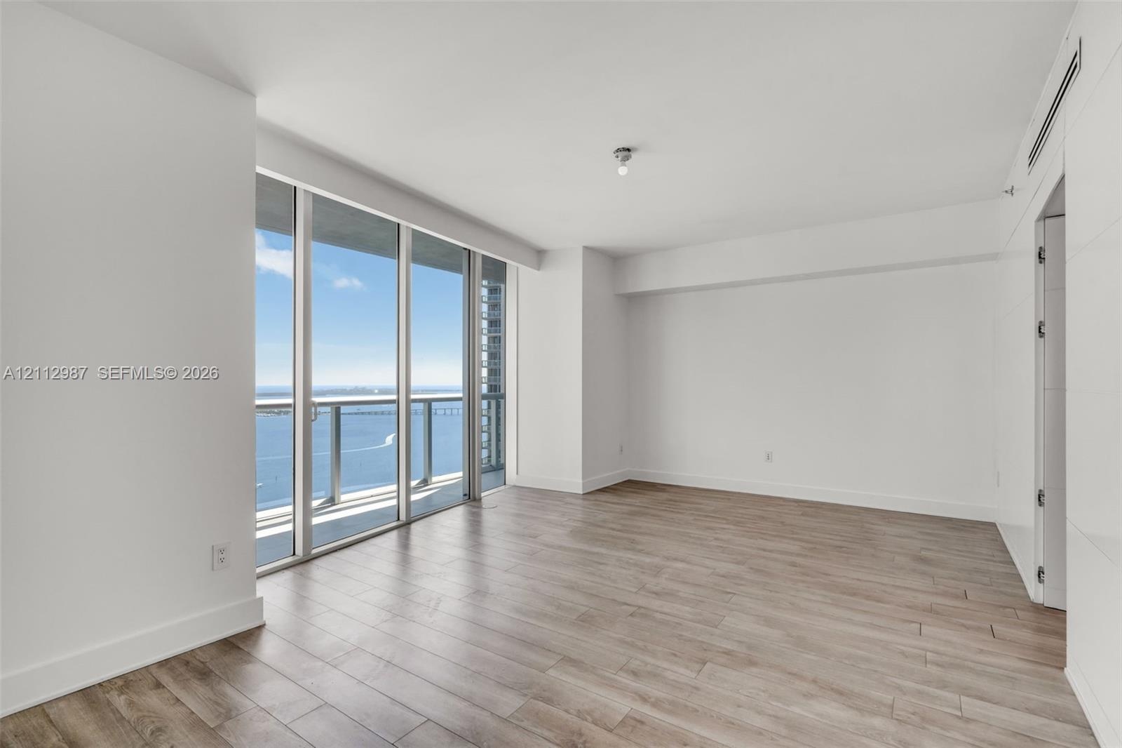 Floor-to-ceiling windows, wood-finish flooring, balcony with glass railing, and water backdrop