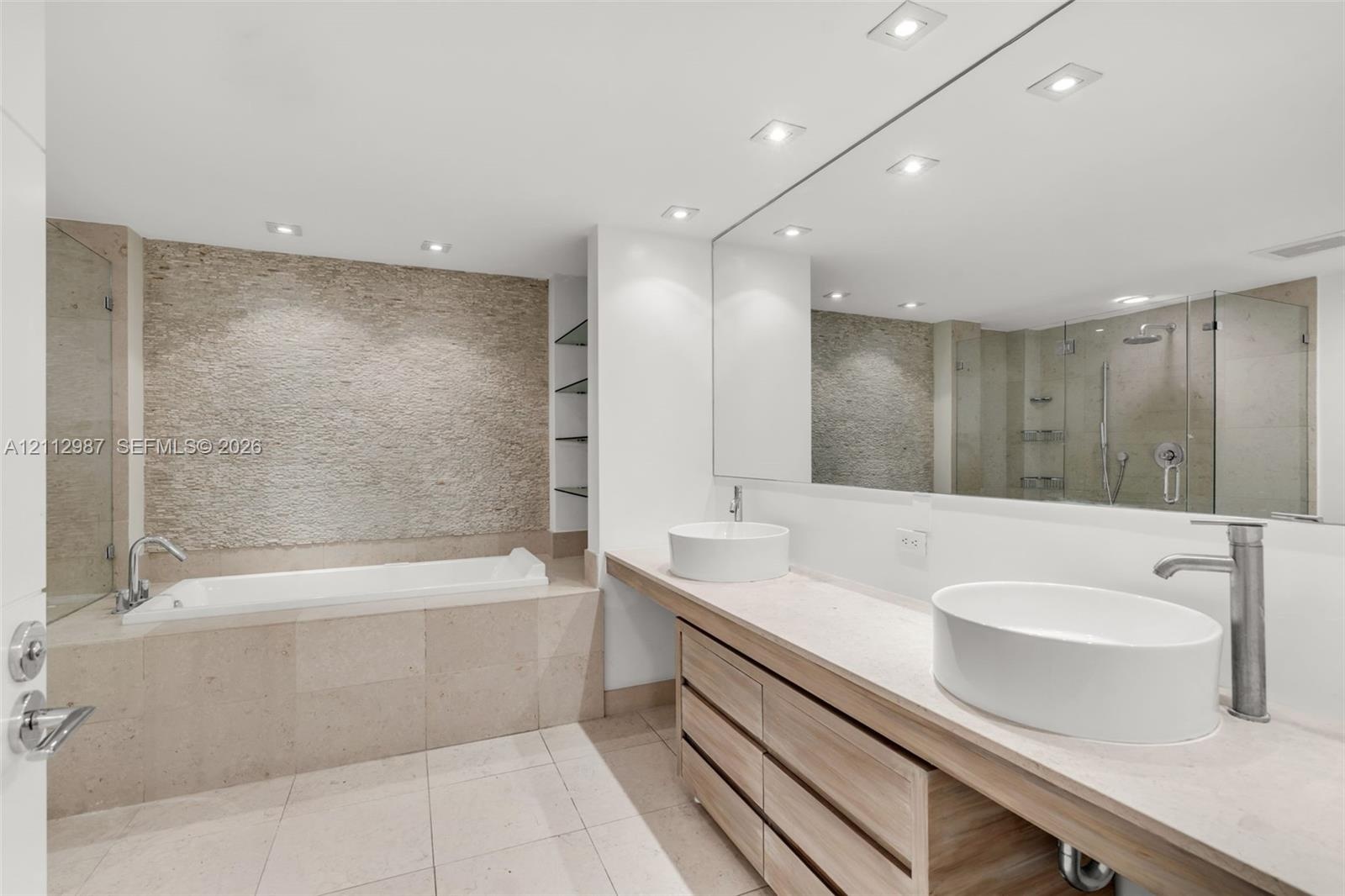 Bathroom featuring dual vessel sinks, a custom vanity with wood-finish cabinetry, a textured stone accent wall, a soaking tub, and a frameless glass s