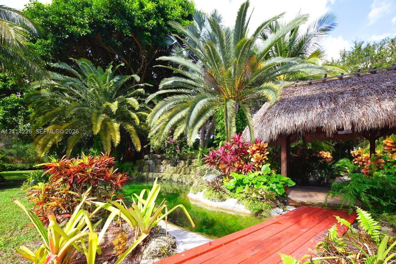 Landscaped garden featuring a thatch-roofed pavilion, a stone-lined pond, mature palms, and a red-finished timber footbridge