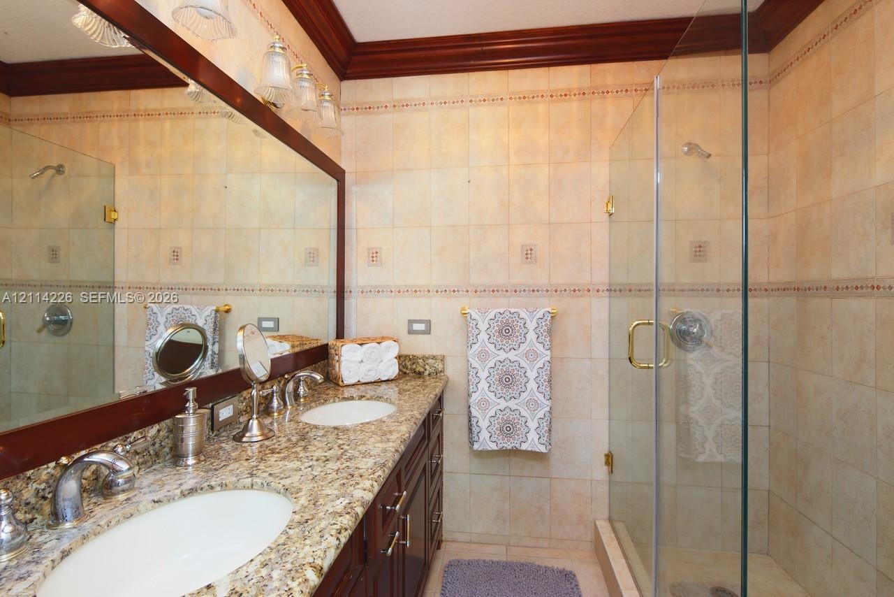 Bathroom featuring a granite countertop vanity with dual undermount sinks, custom dark wood cabinetry, a glass-enclosed shower with tiled walls, and c