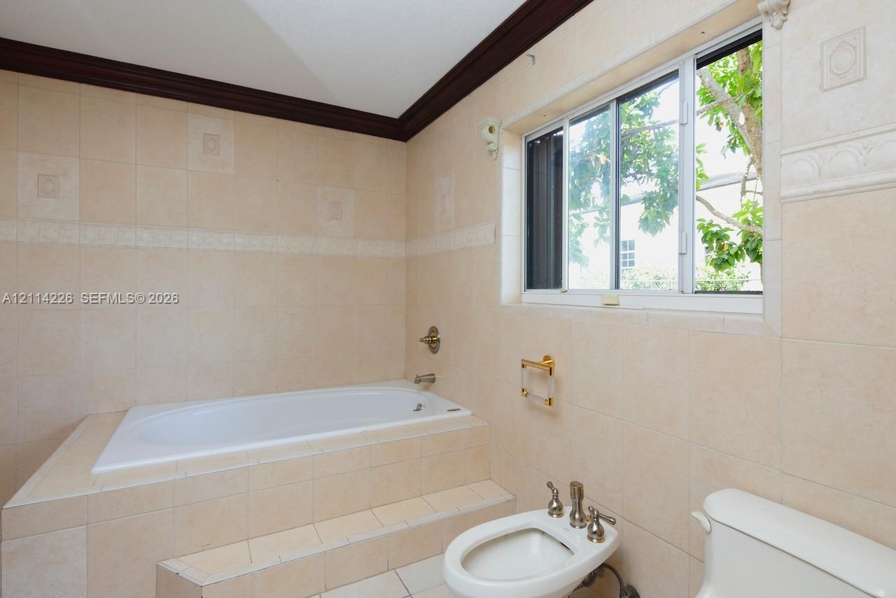 Built-in soaking tub with a tiled surround, neutral ceramic wall tile, dark wood crown molding, sliding window, and a porcelain bidet