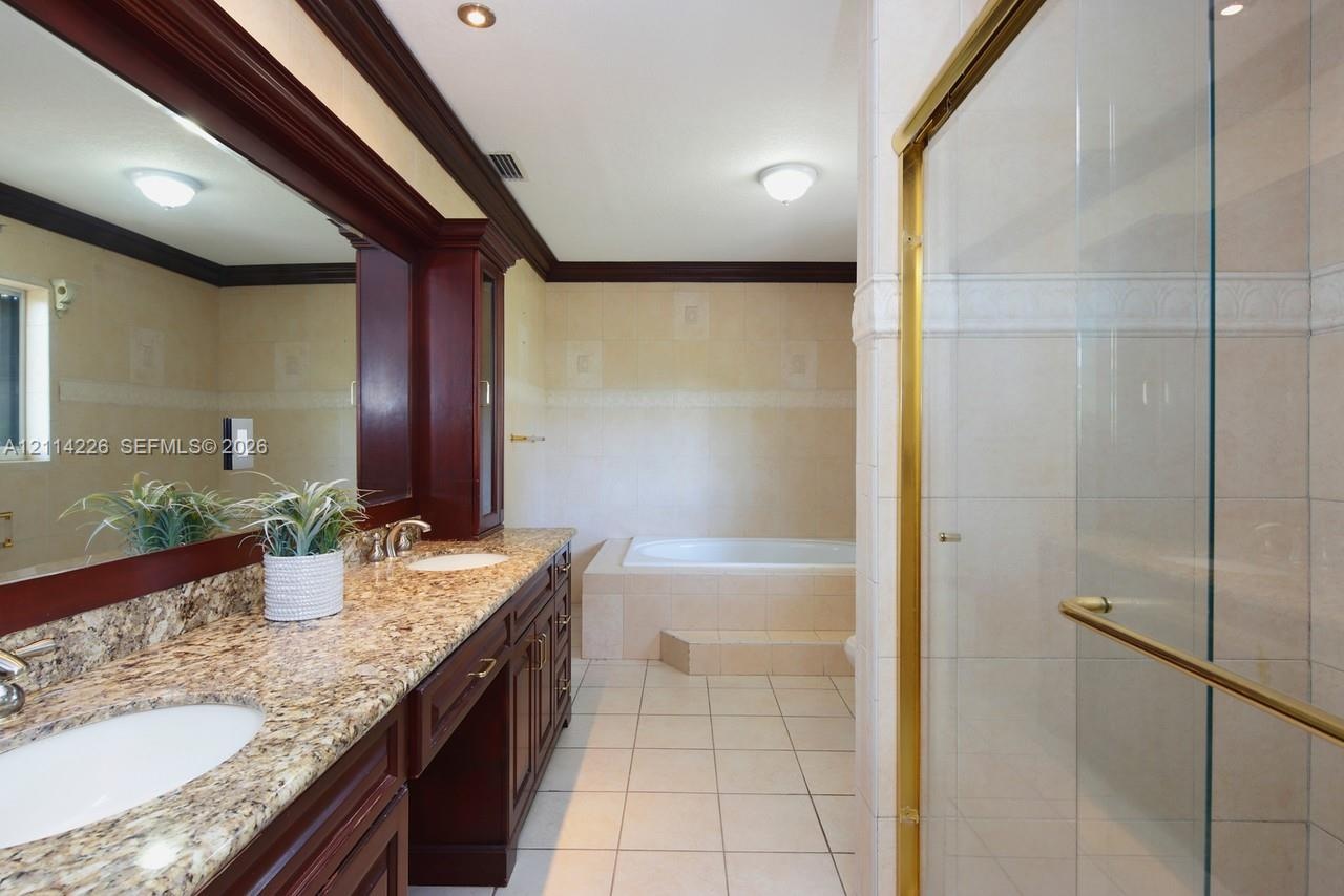 Bathroom featuring a double-sink vanity with dark wood cabinetry and stone-surface countertops, a deep soaking tub with tile surround, a walk-in glass