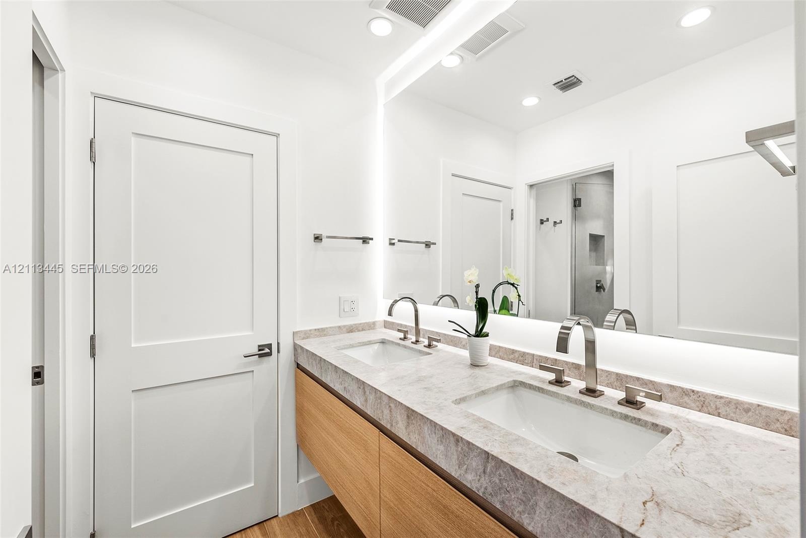 Floating vanity with wood-finish cabinetry, stone countertop, integrated dual undermount sinks, tall arch-neck faucets, and large wall-to-wall mirror 