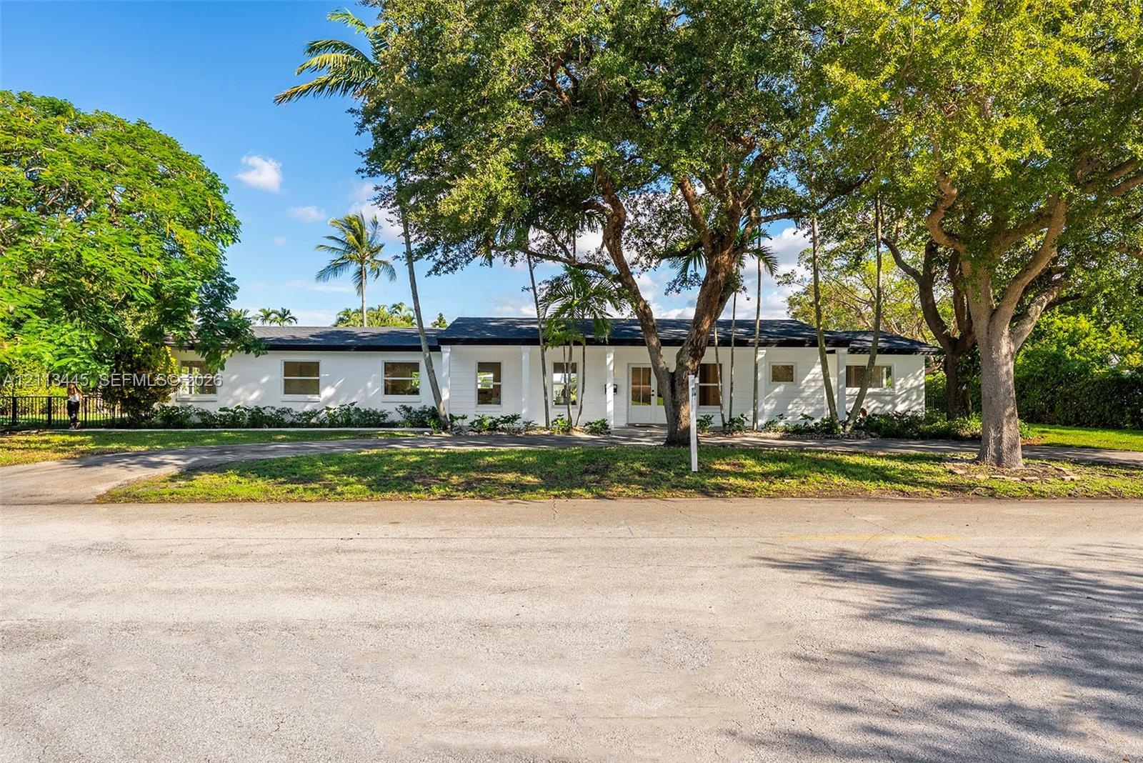 Single-story residence featuring a white exterior, covered front porch, dark shingle roof, paved driveway, and mature landscaping