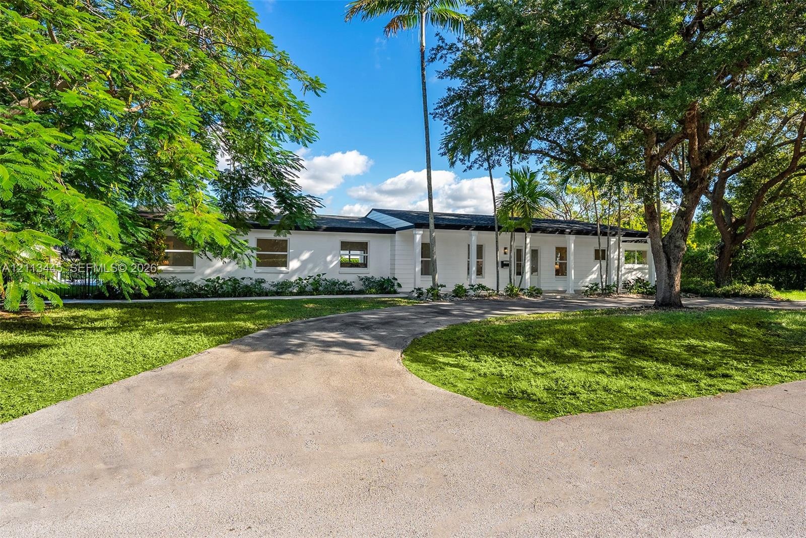 Single-story exterior featuring white masonry walls, a covered front entryway with structural columns, and an expansive paved driveway set within a la
