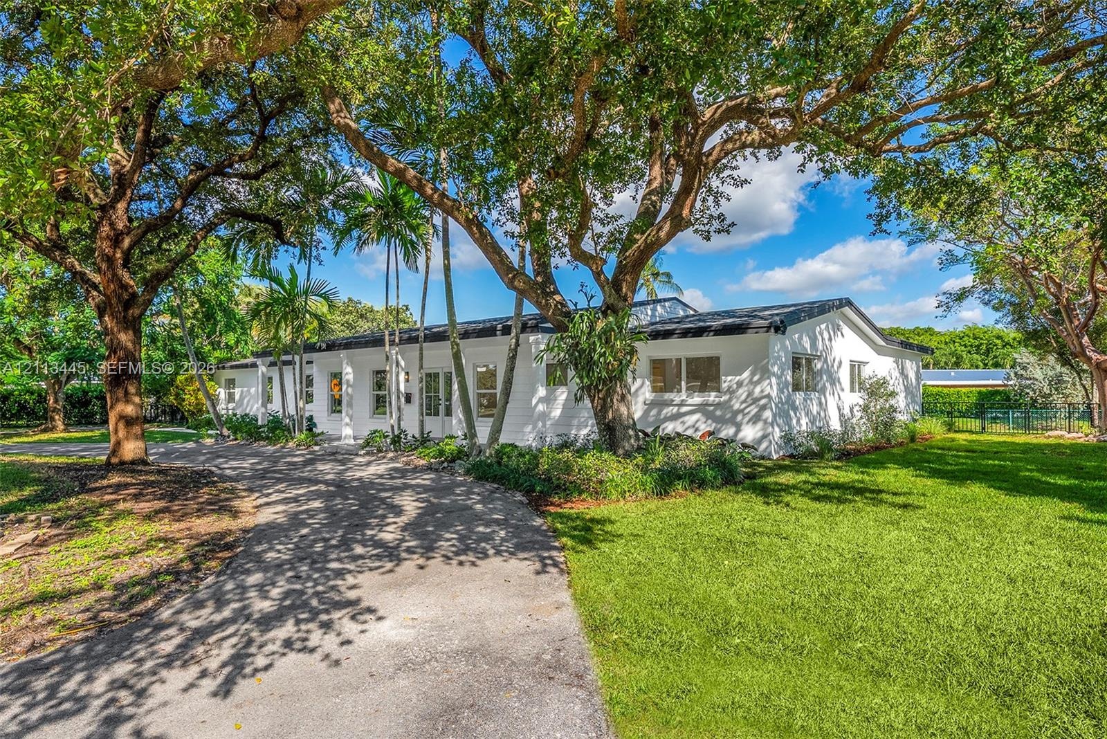 Single-story exterior featuring a white masonry façade, a paved circular driveway, mature oak trees, and palm landscaping