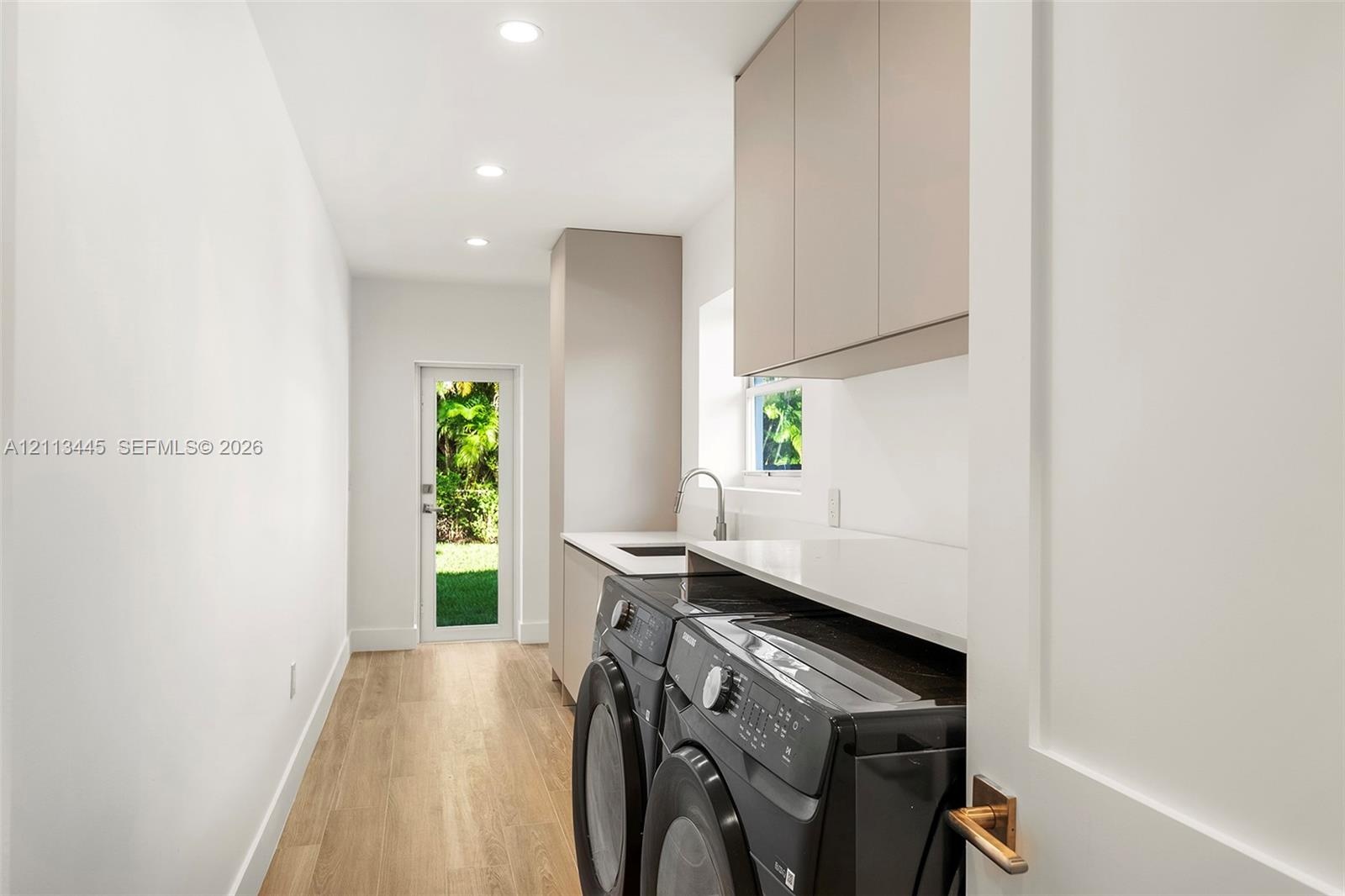 Laundry area featuring minimalist flat-panel cabinetry, light-toned stone-finish countertops, an undermount sink with a gooseneck faucet, wood-finish 