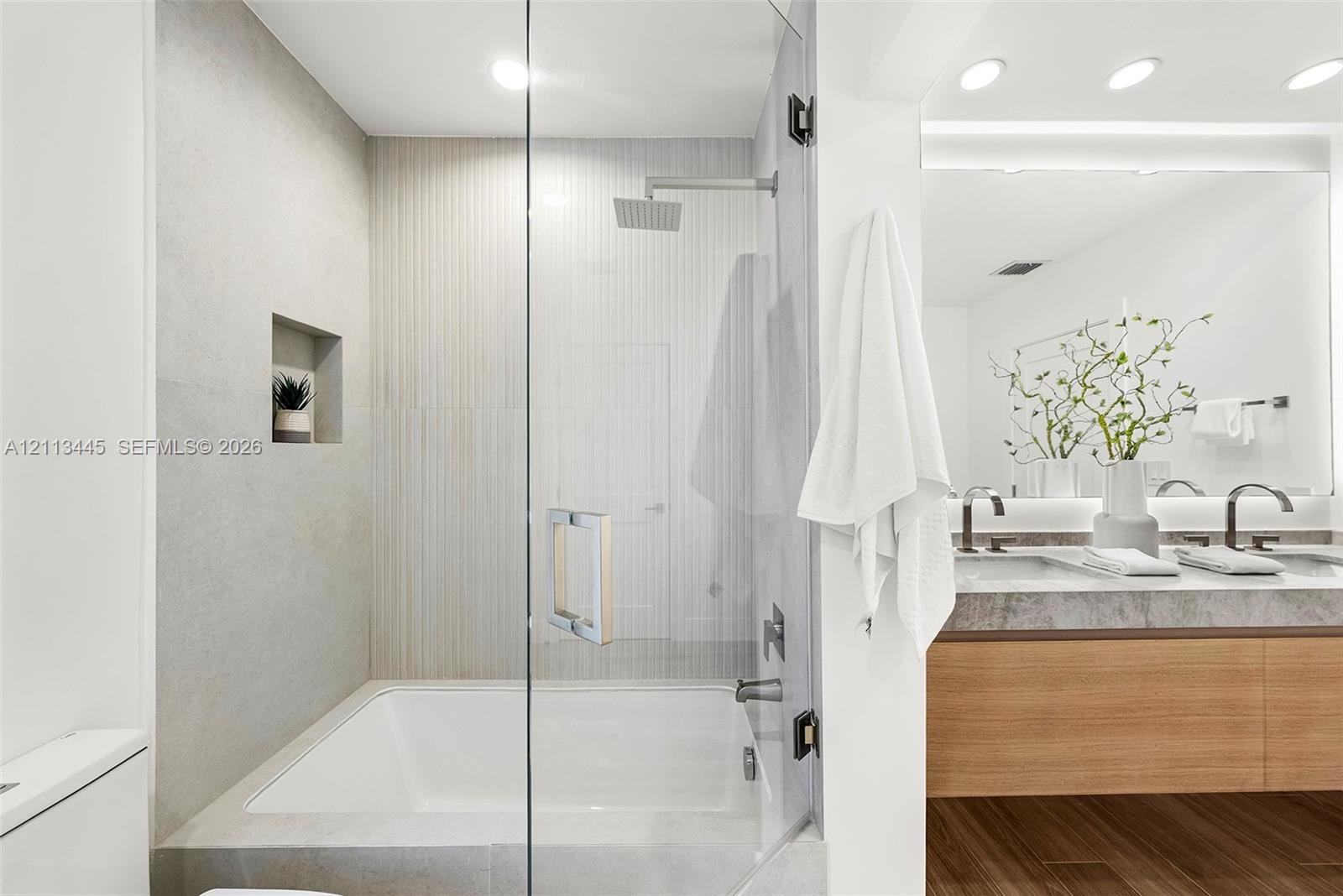 Shower and tub combination featuring glass enclosure, rain-style shower head, built-in recessed wall niche, dual-sink vanity with stone-finish counter