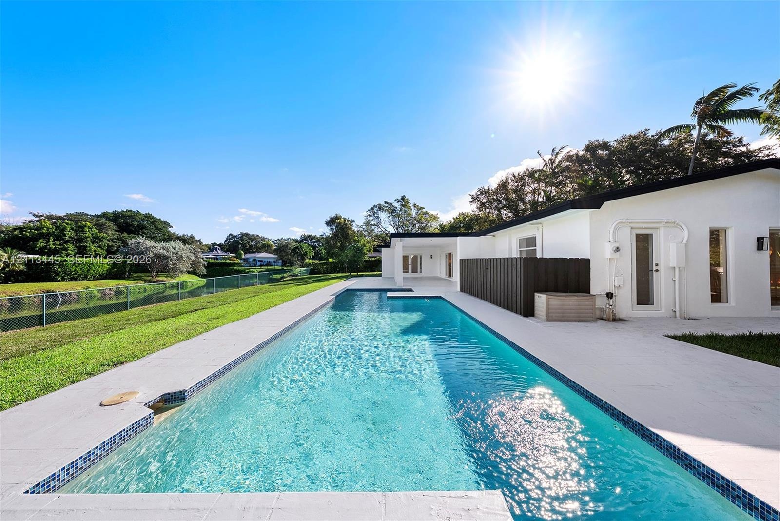 Rectangular in-ground swimming pool with mosaic tile border, light-toned stone pool deck, and manicured grass grounds