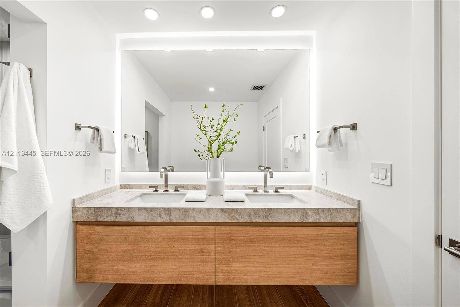Floating wood-finish vanity with dual integrated sinks, stone-surface countertop, LED-backlit mirror, and wall-mounted metal fixtures