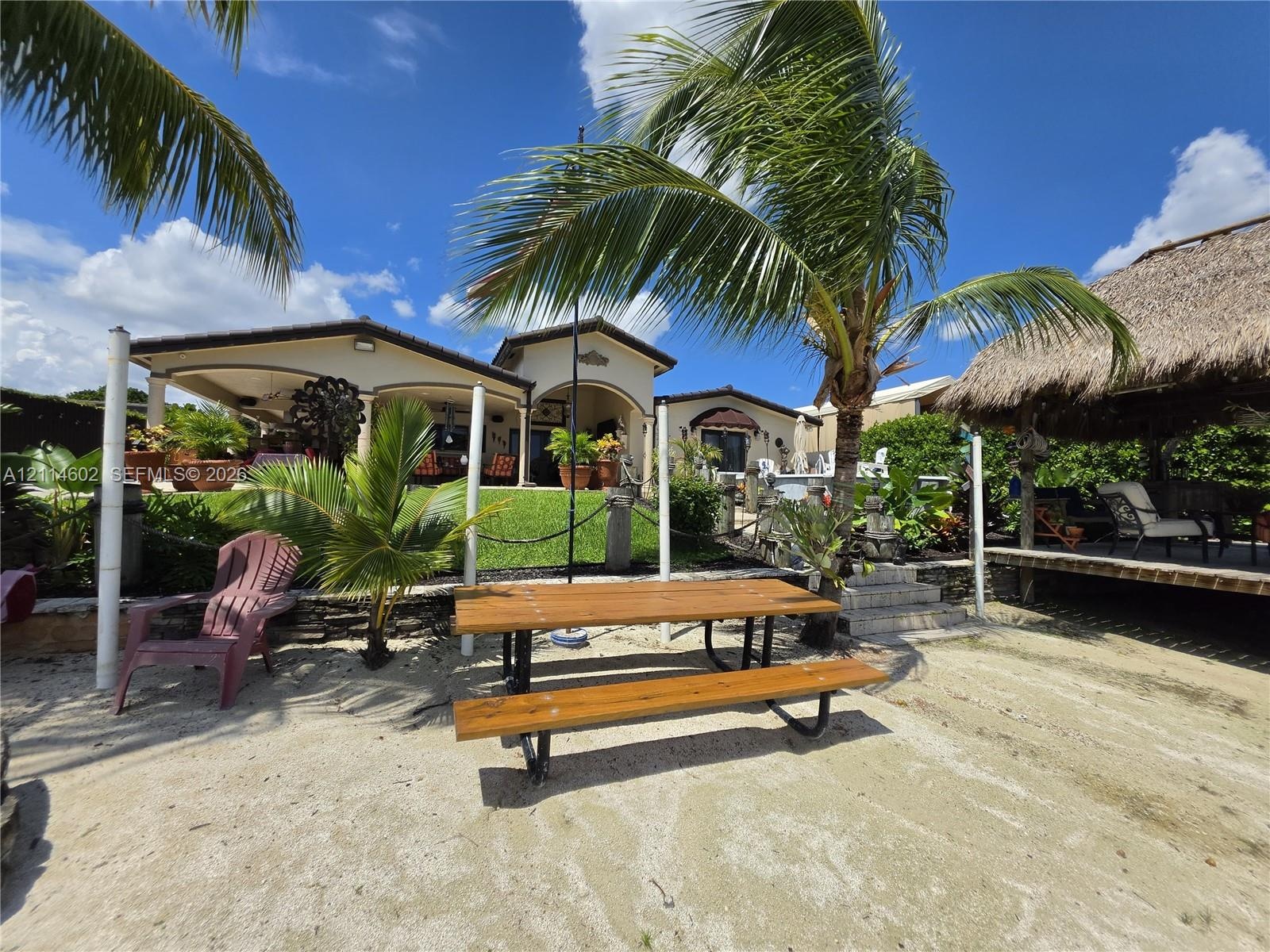 Outdoor living area featuring a covered patio, a thatched-roof tiki hut, sandy shoreline, and mature palm trees