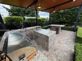 Covered outdoor kitchen featuring stone-clad islands, integrated stainless steel grill, stone-finish countertops, built-in sink, and ceiling fans