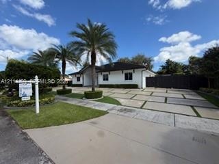 Single-story residence featuring a white exterior, dark shingle roof, expansive paver driveway with turf accents, and mature palm trees
