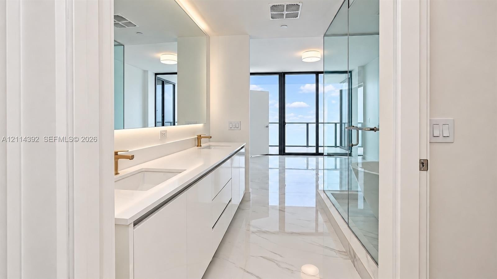 Bathroom featuring white cabinetry, dual undermount sinks with brass-tone fixtures, wall-to-wall mirror, large-format tile flooring, and a glass-enclo