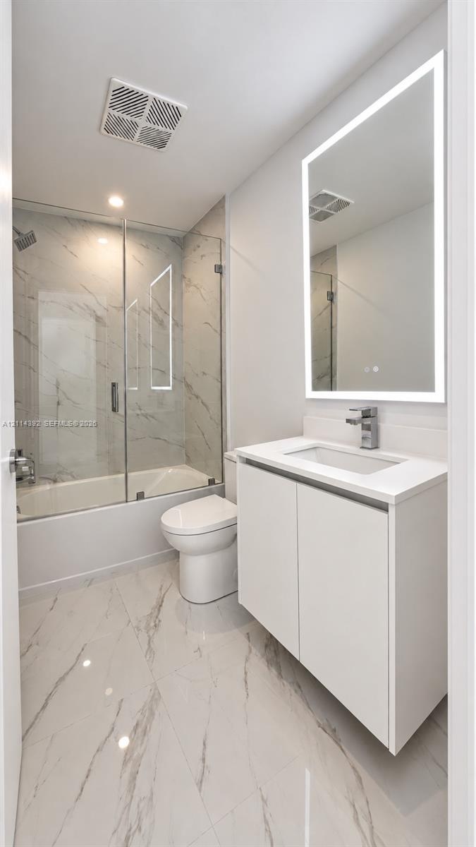 Bathroom featuring a floating vanity with a white countertop, wall-mounted illuminated mirror, porcelain tile flooring, and a glass-enclosed shower wi