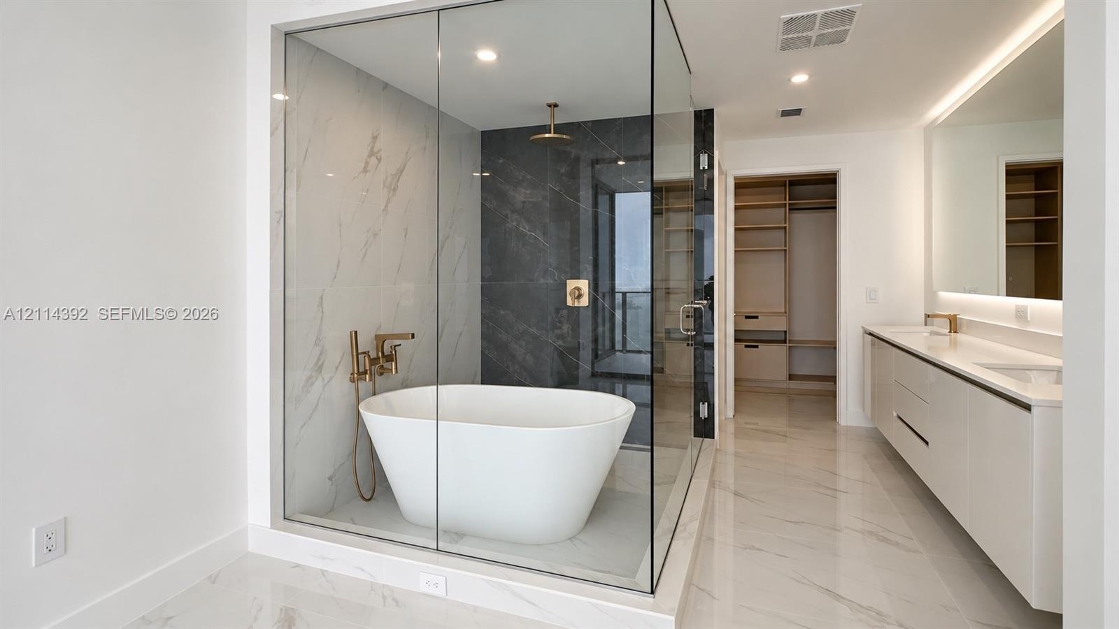Glass-enclosed wet room featuring a freestanding soaking tub, dual-tone tile surround, and brushed gold fixtures, complemented by a dual-sink vanity w