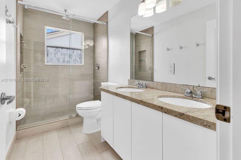 Dual-sink vanity with stone countertop, walk-in shower featuring glass sliding doors and tiled surround, and wood-finish flooring