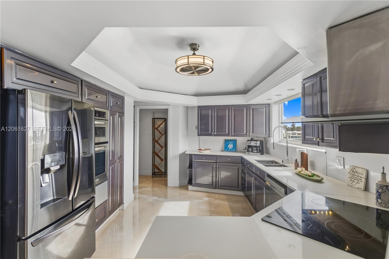 Kitchen featuring dark-toned cabinetry, solid-surface countertops, stainless steel appliances, tray ceiling with recessed lighting, and stone flooring