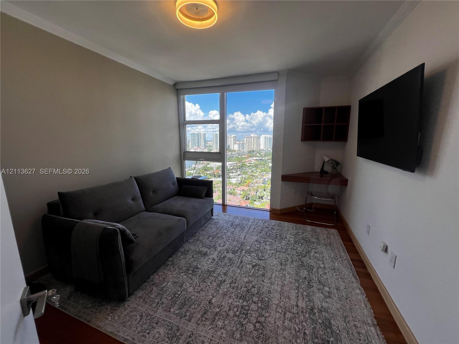 Floor-to-ceiling windows featuring urban scenery, built-in corner desk with integrated shelving, wood-finish flooring, and circular flush-mount ceilin