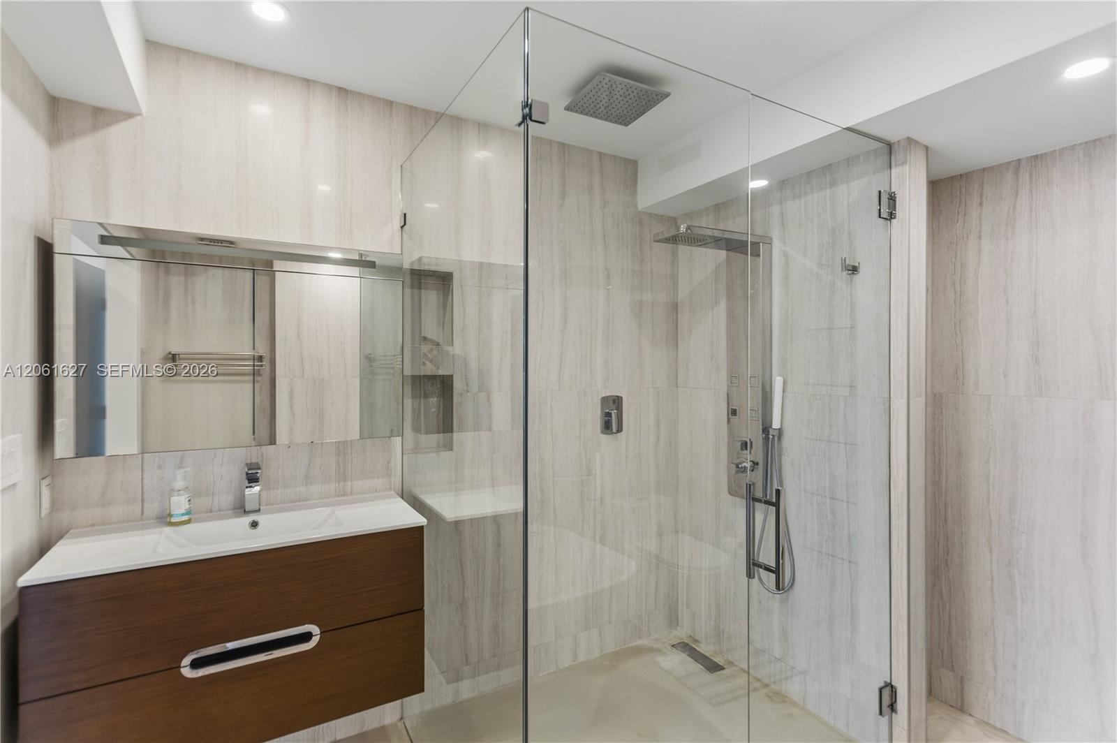 Wall-to-wall stone tile, glass-enclosed shower featuring a rainfall showerhead and integrated body sprays, floating wood-finish vanity, and integrated