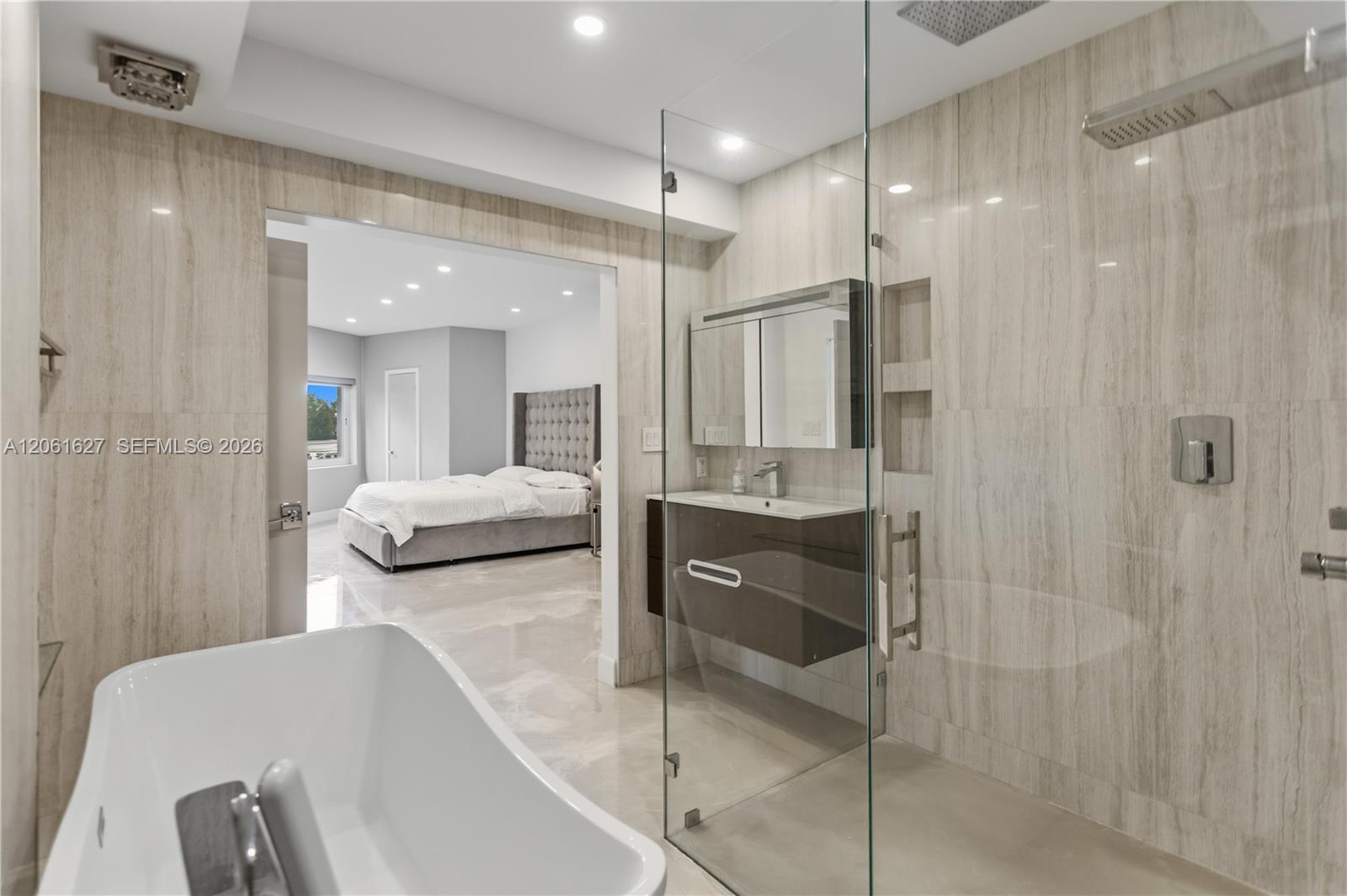 Full-height stone-tiled walls, freestanding soaking tub, glass-enclosed walk-in shower with rainfall fixture, recessed shower niche, and wall-mounted 
