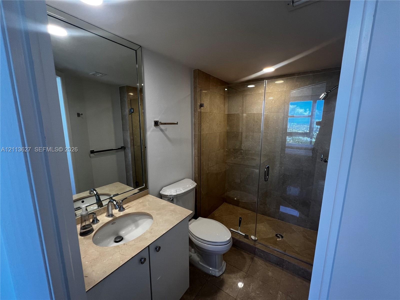 Bathroom featuring a stone-surface vanity with an integrated basin, large wall-mounted mirror, and a walk-in shower with stone tile surround and glass