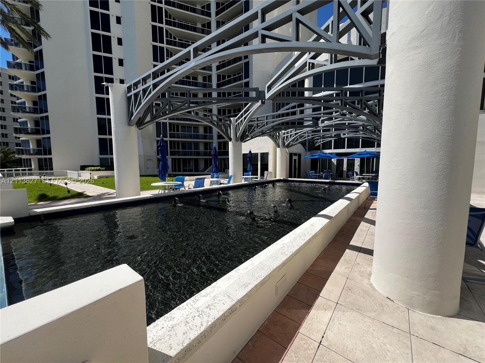Rectangular reflective pool with built-in water features, architectural overhead metal framing, cylindrical support columns, and light-toned stone til