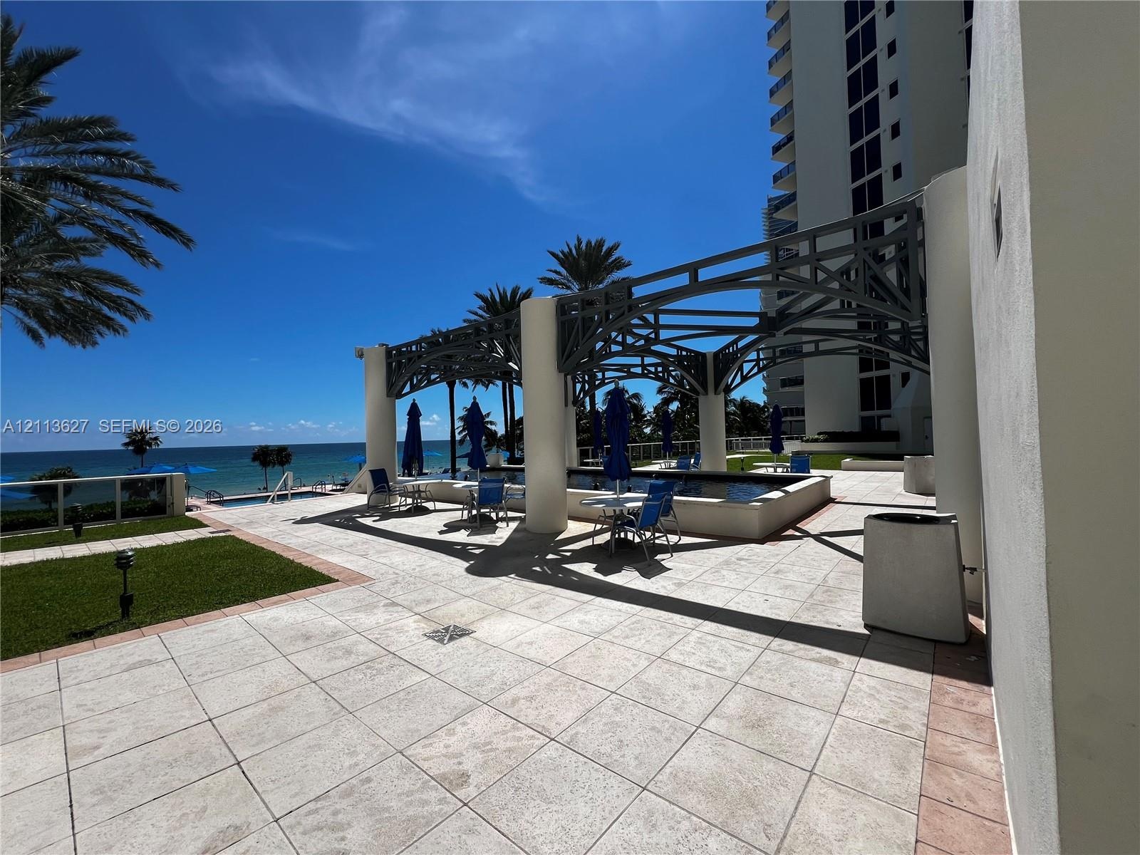 Outdoor terrace featuring stone tile flooring, a structured metal pergola, swimming pool, manicured lawn, and ocean backdrop