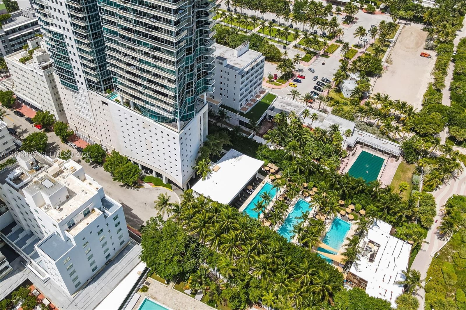 High-rise residential structure featuring glass-paneled balconies, multiple swimming pools, extensive lush landscaping with mature palm trees, and har