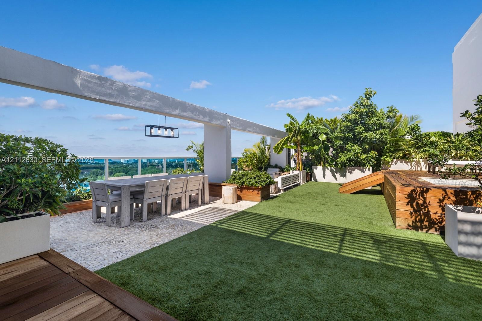 Rooftop terrace featuring synthetic lawn, wood-decking accents, stone-tile flooring, glass panel railing, and overhead structural beam with integrated