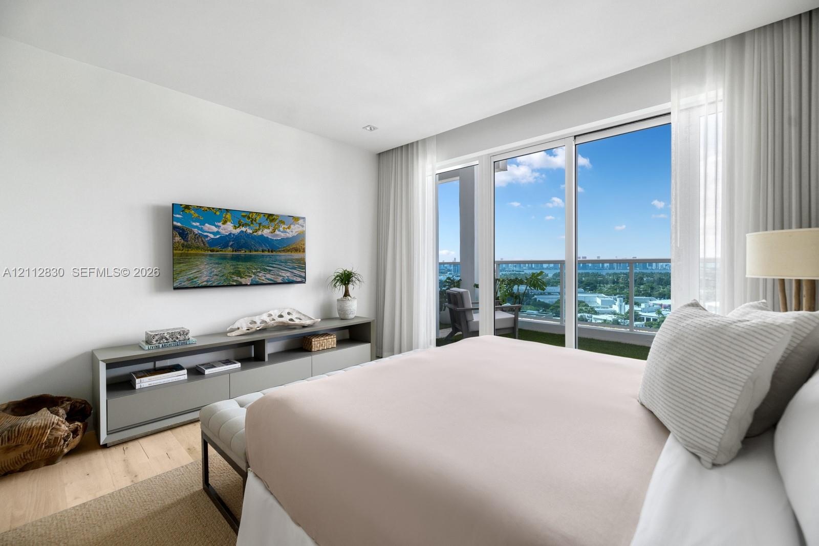 Floor-to-ceiling sliding glass doors, built-in gray media console, light wood-finish flooring, and balcony with metal railing