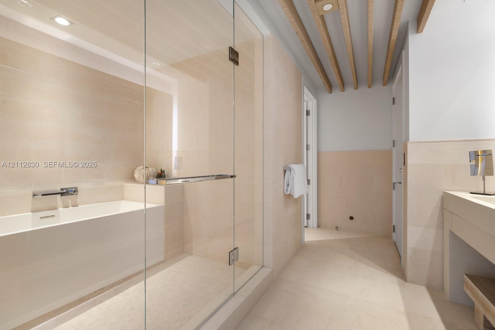 Bathroom featuring a glass-enclosed shower, soaking tub with wall-mounted faucet, neutral stone-finish wall and floor tile, and exposed wooden ceiling