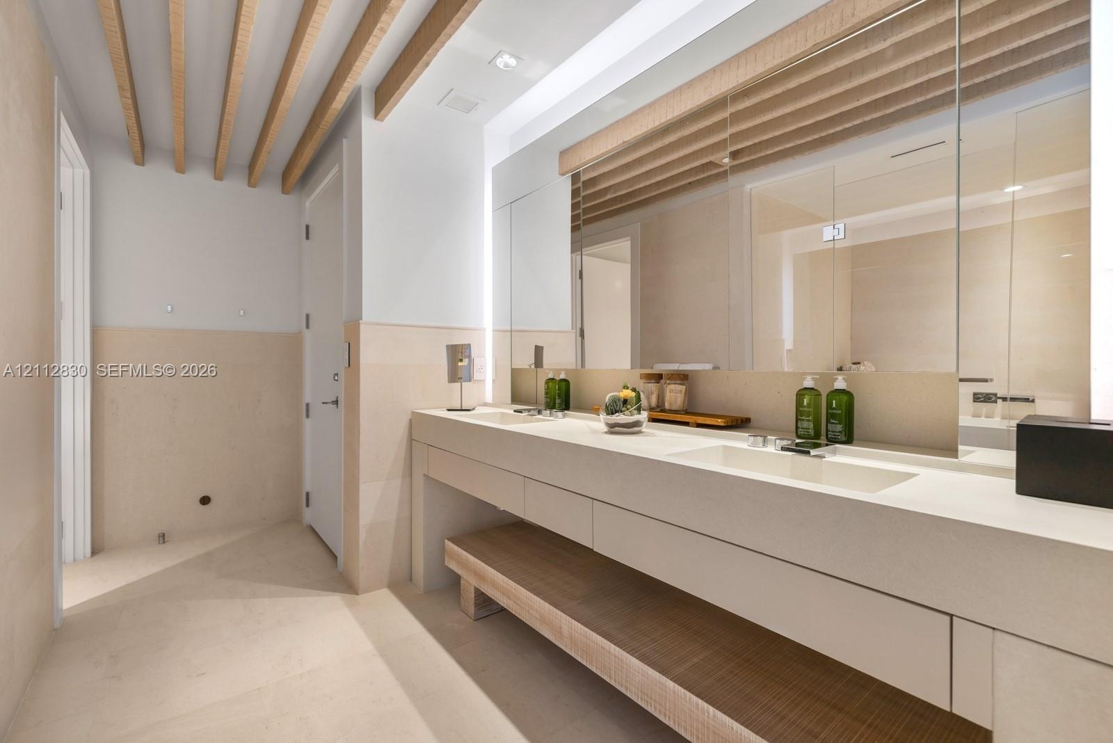 Dual vanity with integrated sinks, stone-surface countertop, full-width mirror with integrated lighting, exposed wood ceiling beams, and stone-finish 