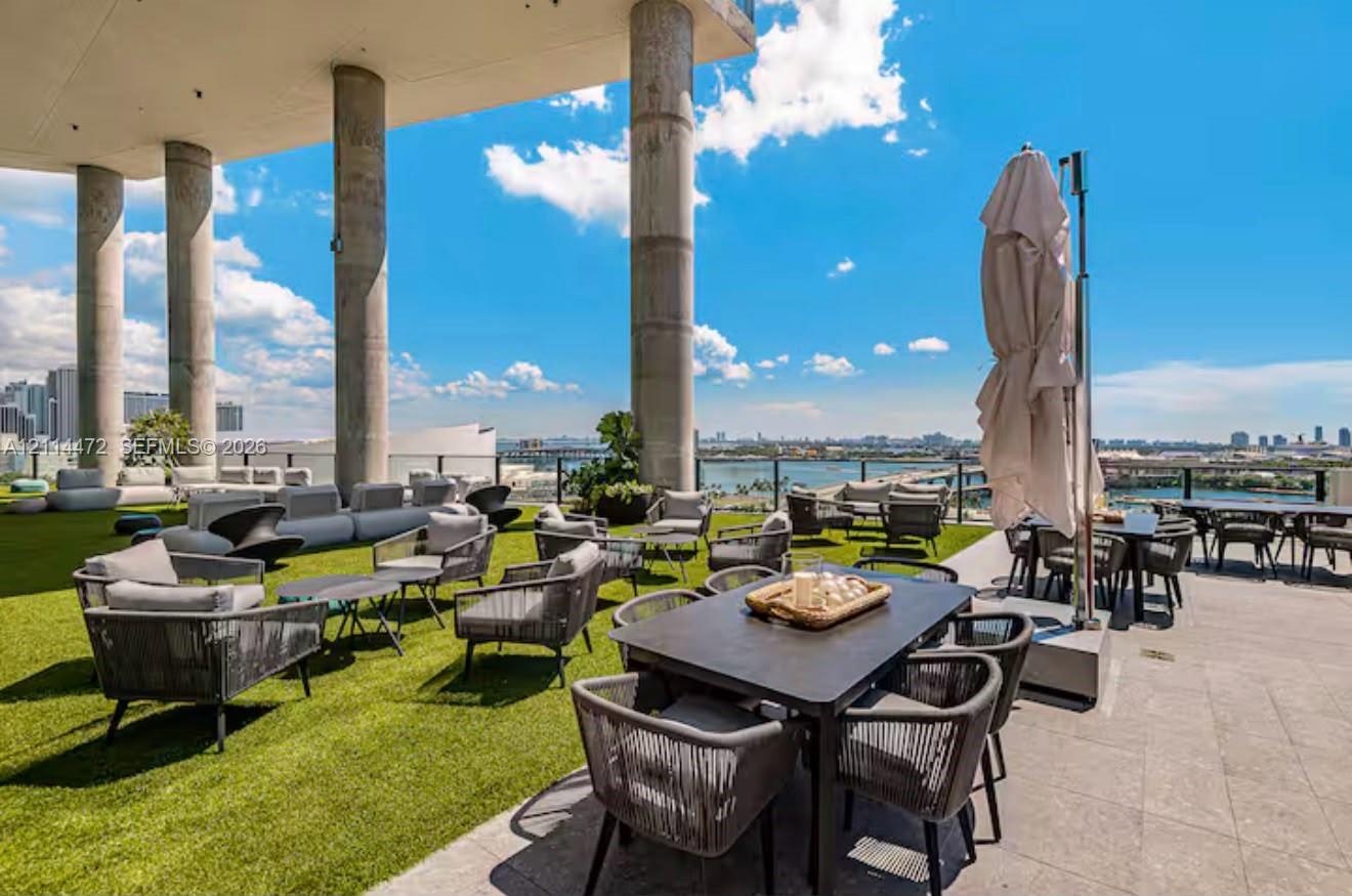 Expansive rooftop terrace featuring synthetic turf flooring, stone tile decking, structural concrete columns, and a glass panel perimeter railing