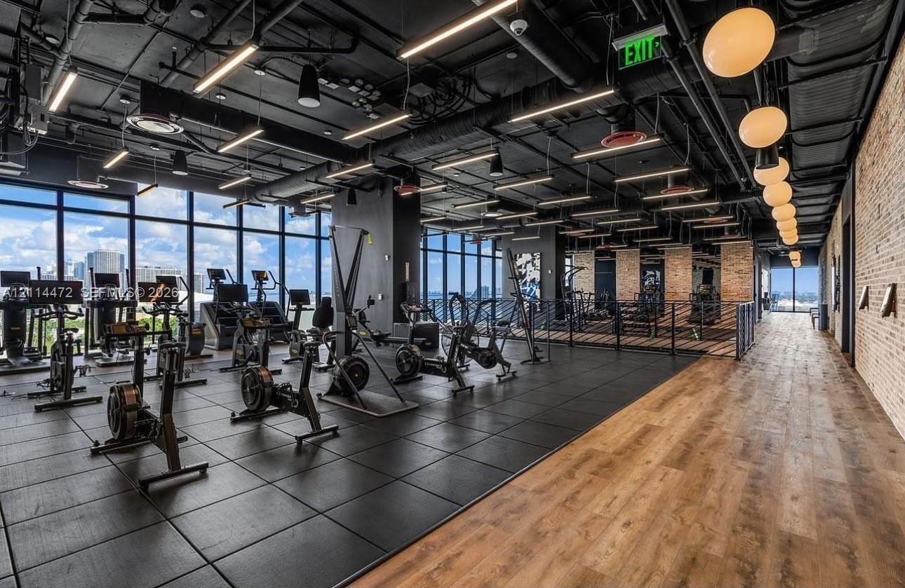 Fitness center featuring floor-to-ceiling windows, exposed industrial ceilings with recessed lighting, wood-finish flooring, and a textured brick acce