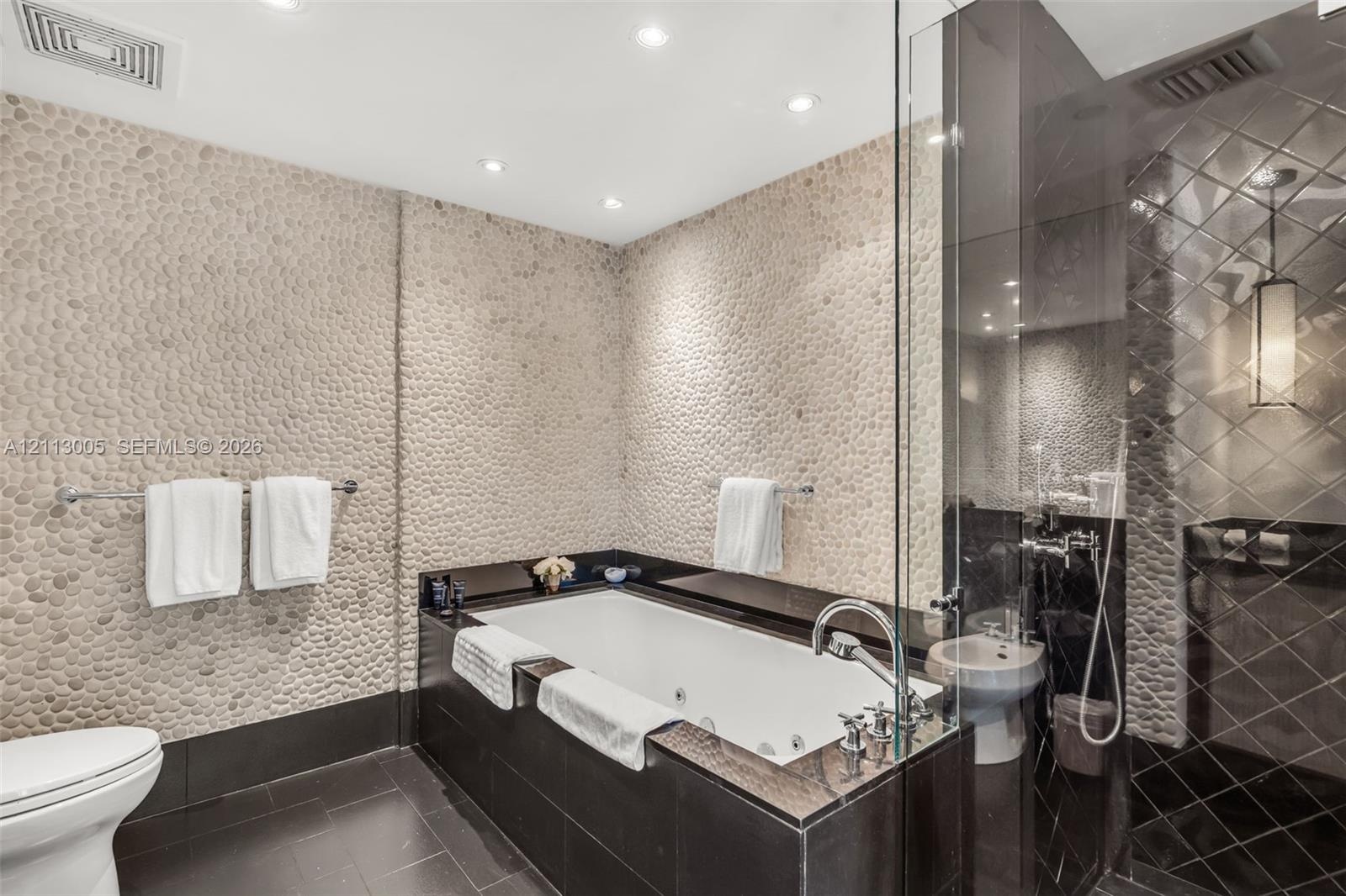 Bathroom featuring pebble-textured stone accent walls, a jetted soaking tub with a dark stone surround, a glass-enclosed shower with diamond-patterned