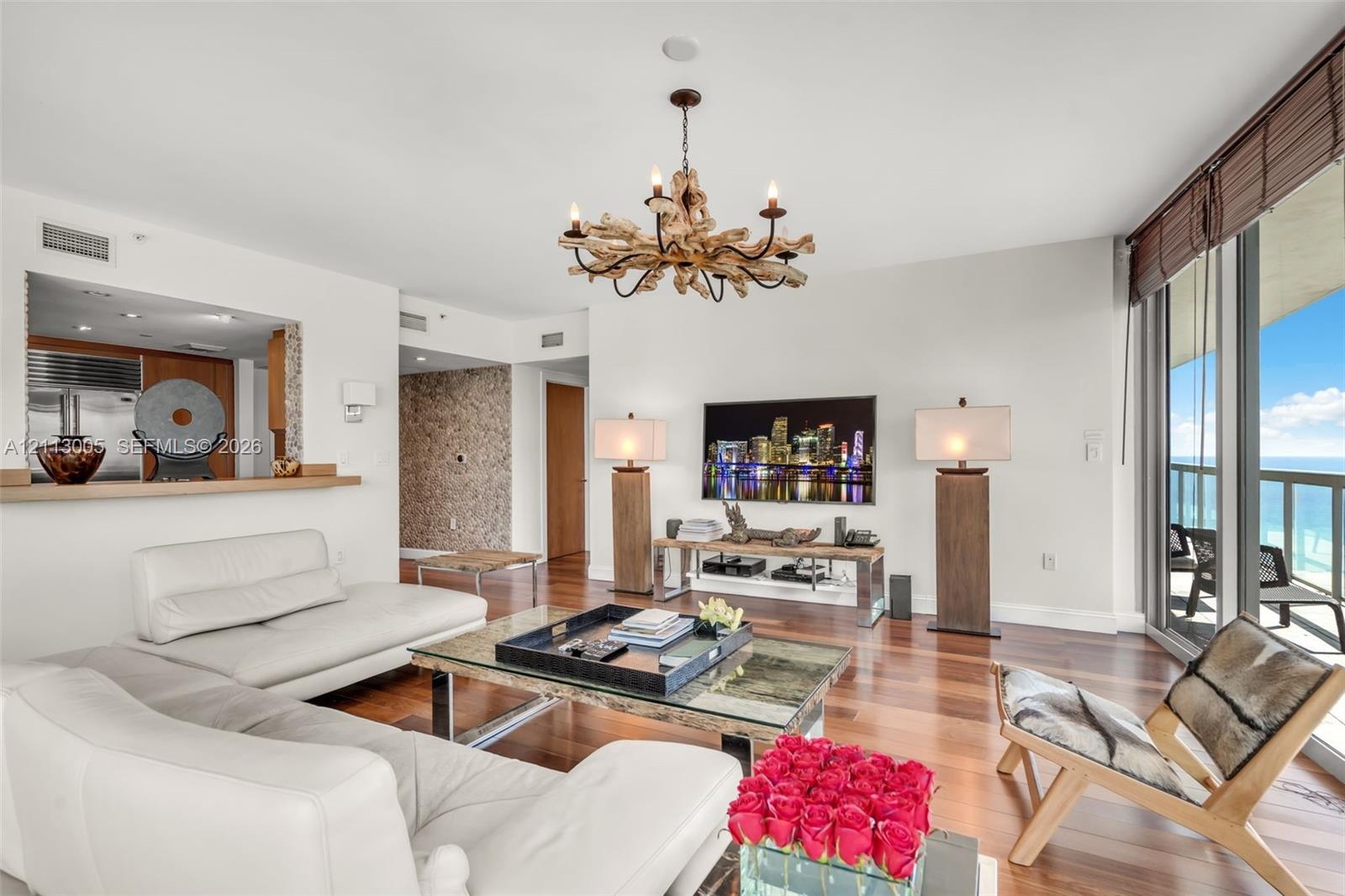 Living area featuring wood-finish flooring, a textured stone feature wall, a custom driftwood-style chandelier, and floor-to-ceiling windows with balc
