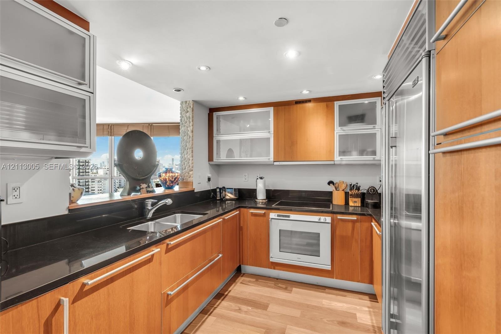 Kitchen featuring wood-veneer cabinetry with frosted glass inserts, dark solid-surface countertops, built-in stainless steel refrigerator, recessed li