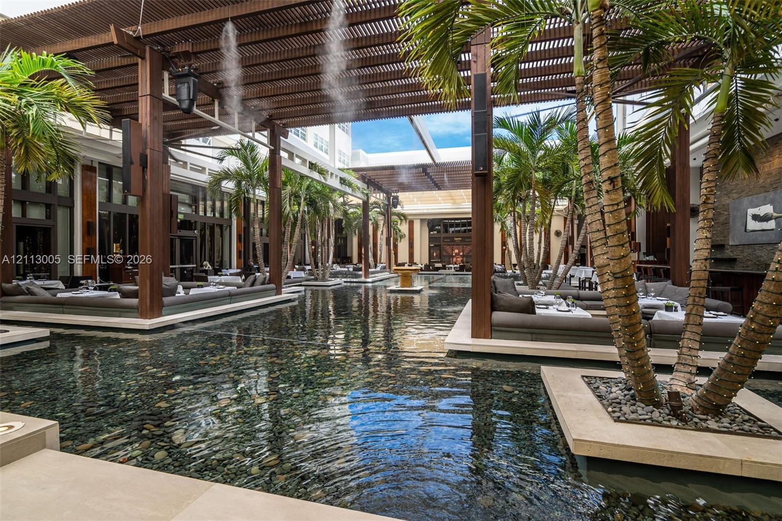 Expansive courtyard featuring a reflection pool, overhead wood-slat pergola, integrated palm tree planters, stone tile hardscaping, and built-in mason