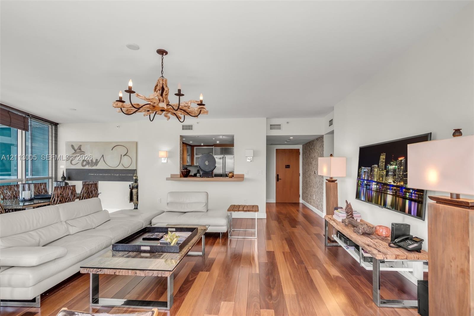 Open-concept living area featuring wood-finish flooring, a natural driftwood chandelier, textured wall accents, and a service pass-through window with