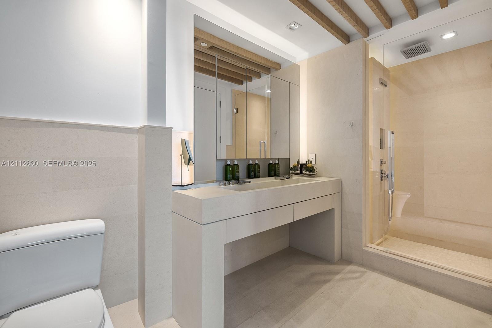 Stone-surfaced floating vanity, ceiling with exposed wood-finish beams, integrated medicine cabinet, and glass-enclosed walk-in shower with stone tile