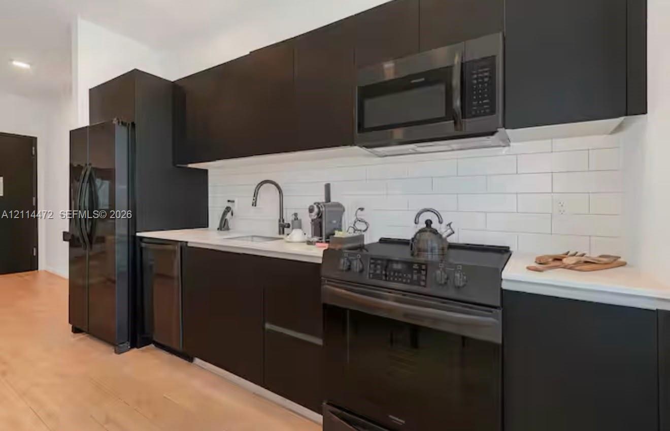 Modern kitchen featuring matte black flat-panel cabinetry, white solid surface countertops, white subway tile backsplash, and a suite of black applian