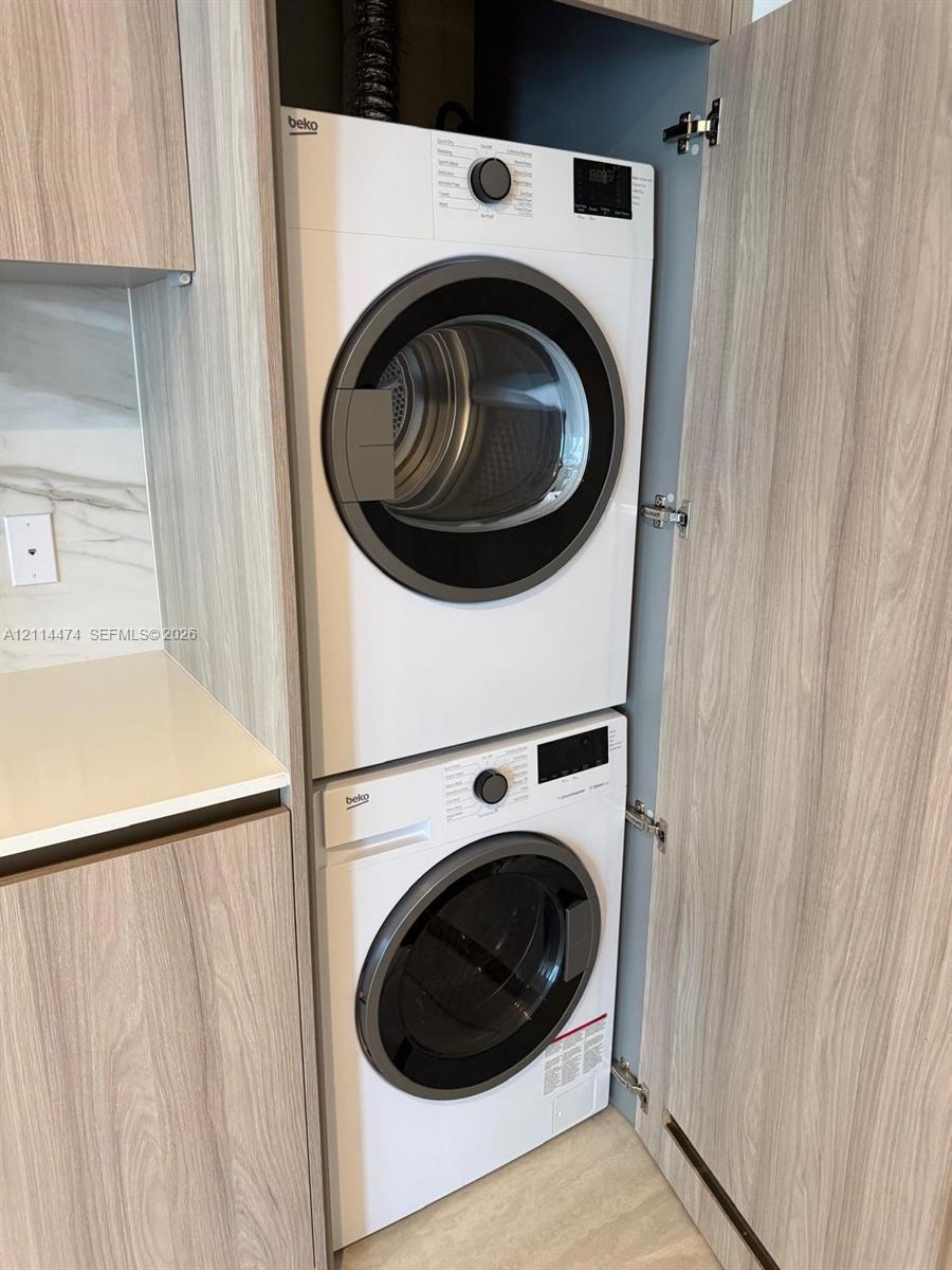 Built-in stacked laundry unit with light-toned wood-finish cabinetry, quartz countertop, and stone-finish backsplash
