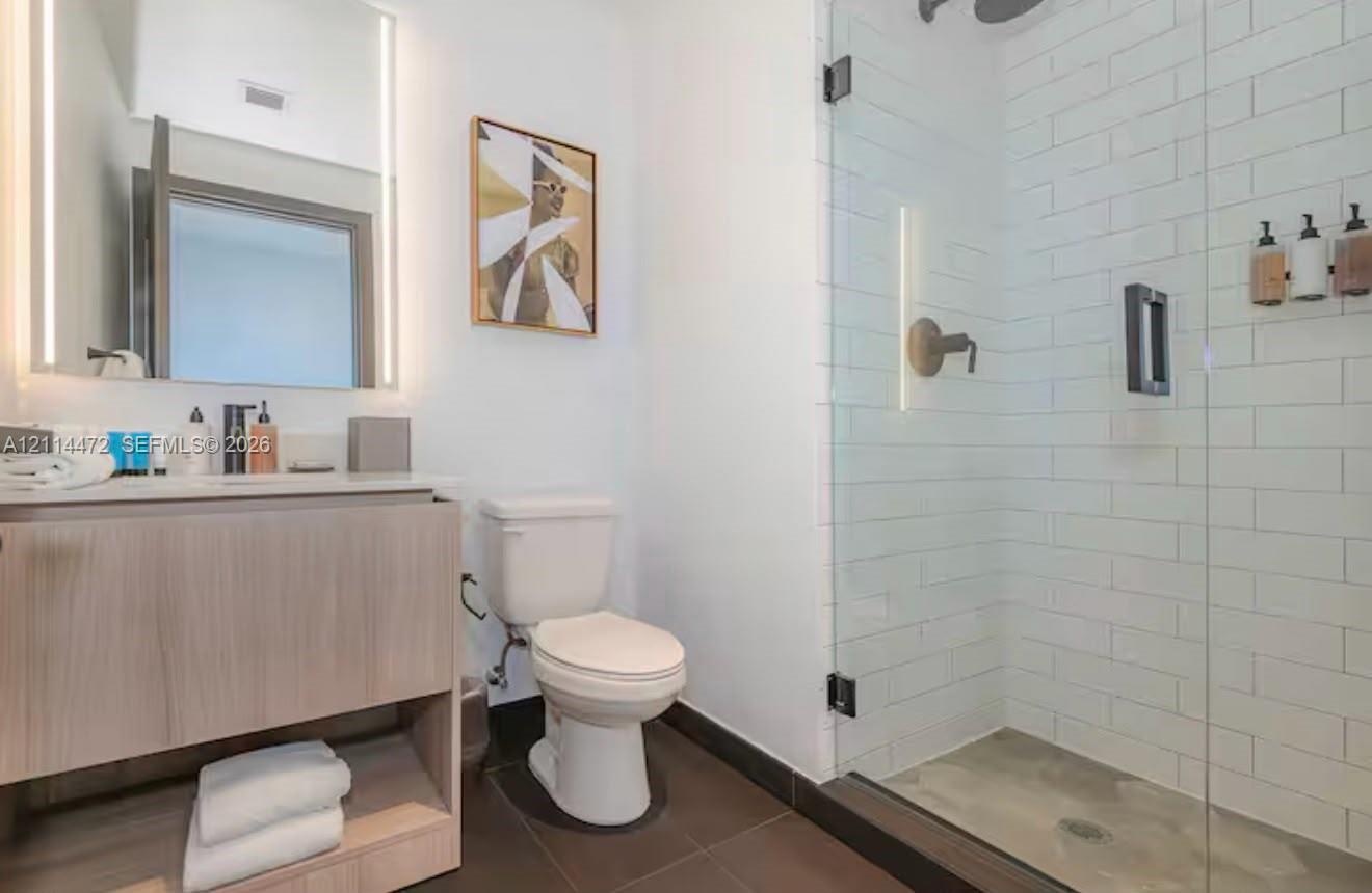 Wood-finish floating vanity with an integrated countertop, illuminated wall-mounted mirror, walk-in shower with white subway tile surround, and large-